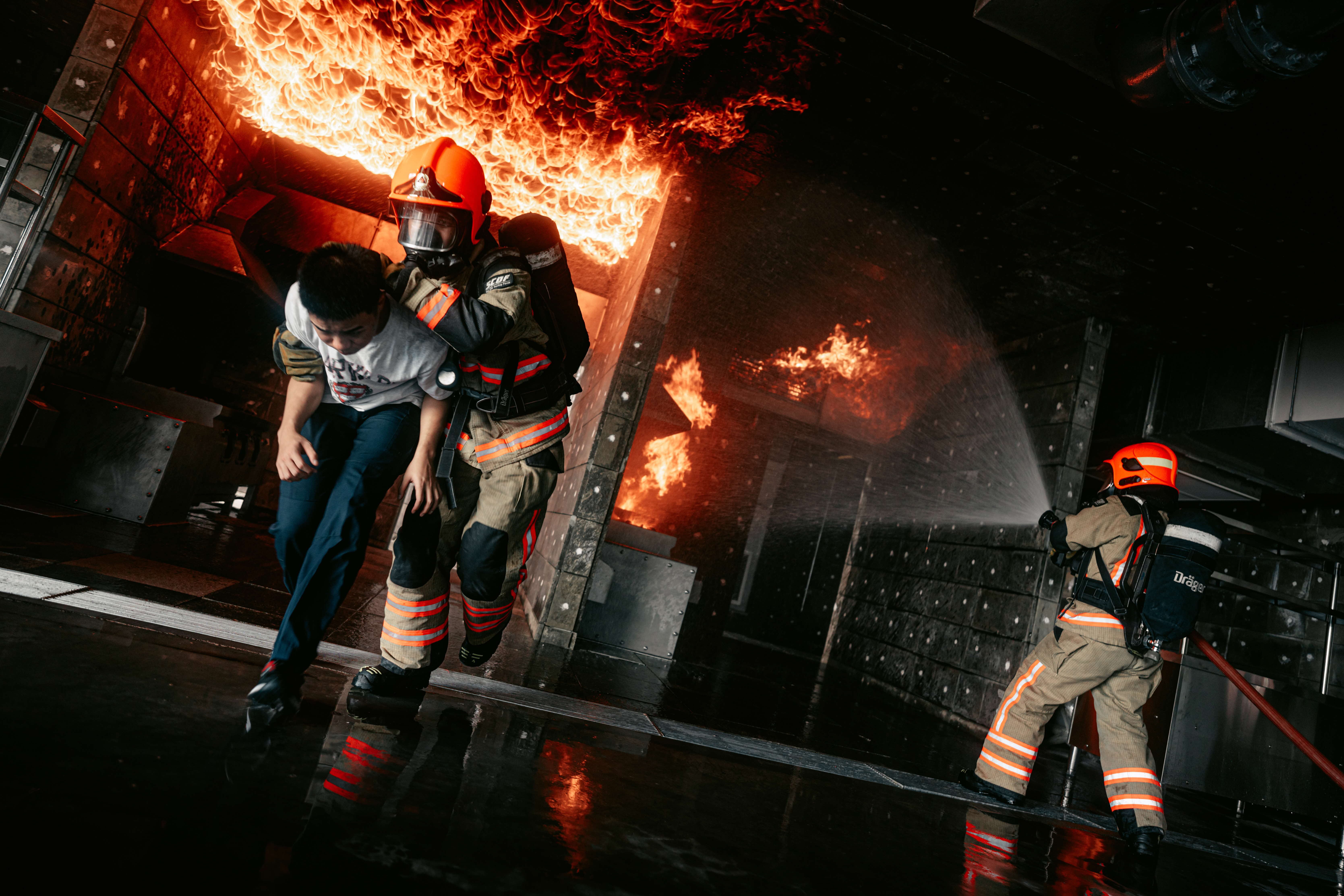 Firefighter carrying person from fire, another firefighter sprays water with Dräger tank.